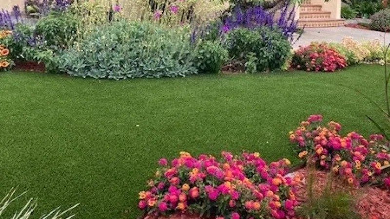 Front yard with vibrant flower beds, lush green lawn, and tropical plants surrounding a beige house with arches.