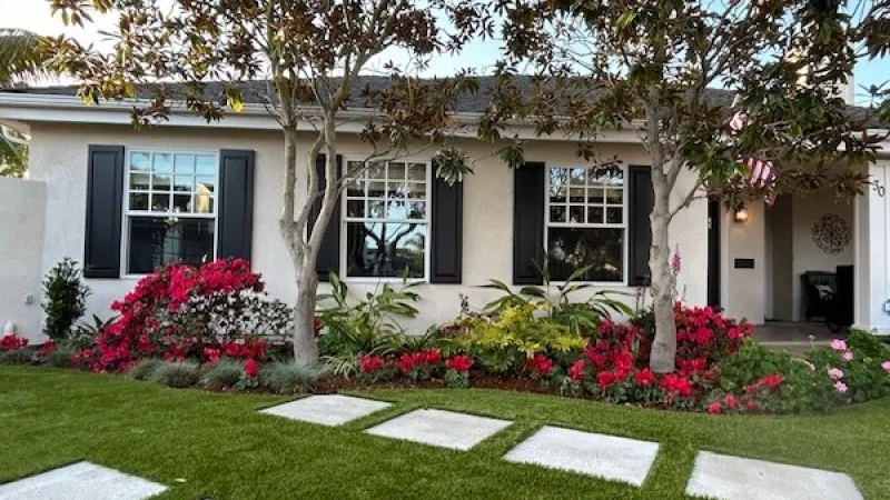 Front yard with a well-maintained lawn, stepping stones, colorful flower beds, and a house with black shutters.