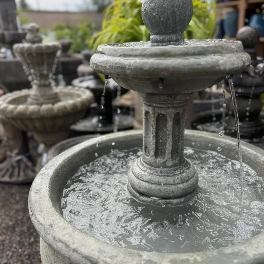 Close-up of a decorative stone garden fountain with flowing water and blurred background plants and pots.