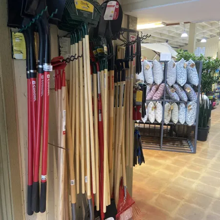 Display of shovels, rakes, and garden tools with decorative pillows on shelves in a store aisle.
