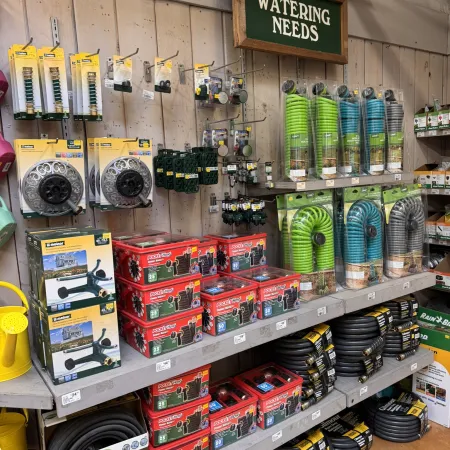 Shelf display of various garden watering tools and hoses under a wooden sign labeled Watering Needs.