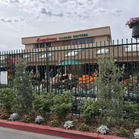 Arroyo Garden Center entrance with plants, pumpkins, and customers behind a metal fence under cloudy sky