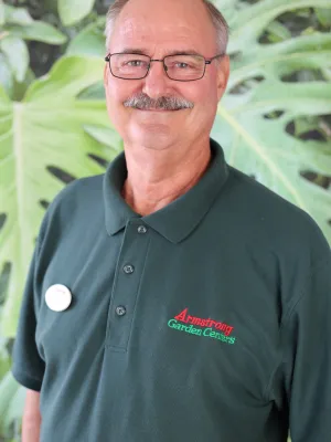 Smiling man with glasses wearing a dark green Armstrong Garden Centers polo outdoors with large green leaves in background