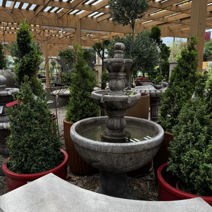 Stone water fountain surrounded by potted topiary trees under a wooden pergola in a garden center.