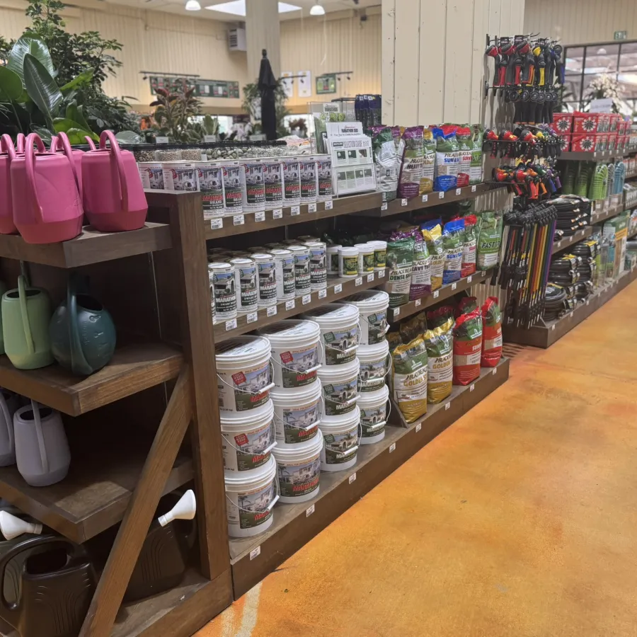 Garden store aisle with shelves of watering cans, plant food, gardening tools, and various supplies on display.
