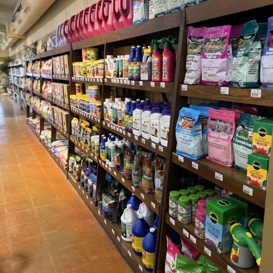 Aisle of gardening supplies including fertilizers, plant food, watering cans, and pest control products on wooden shelves.