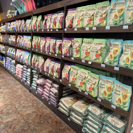 Wooden shelves stocked with various seed packets and gardening supplies in a store aisle with tiled floor.