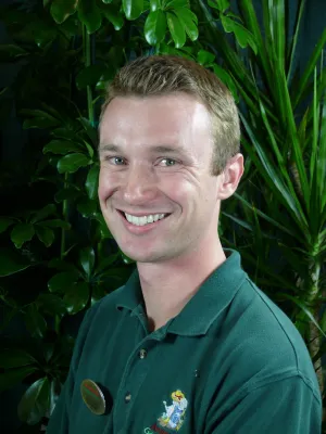 Smiling man wearing green polo shirt with embroidered logo standing in front of leafy green plants.