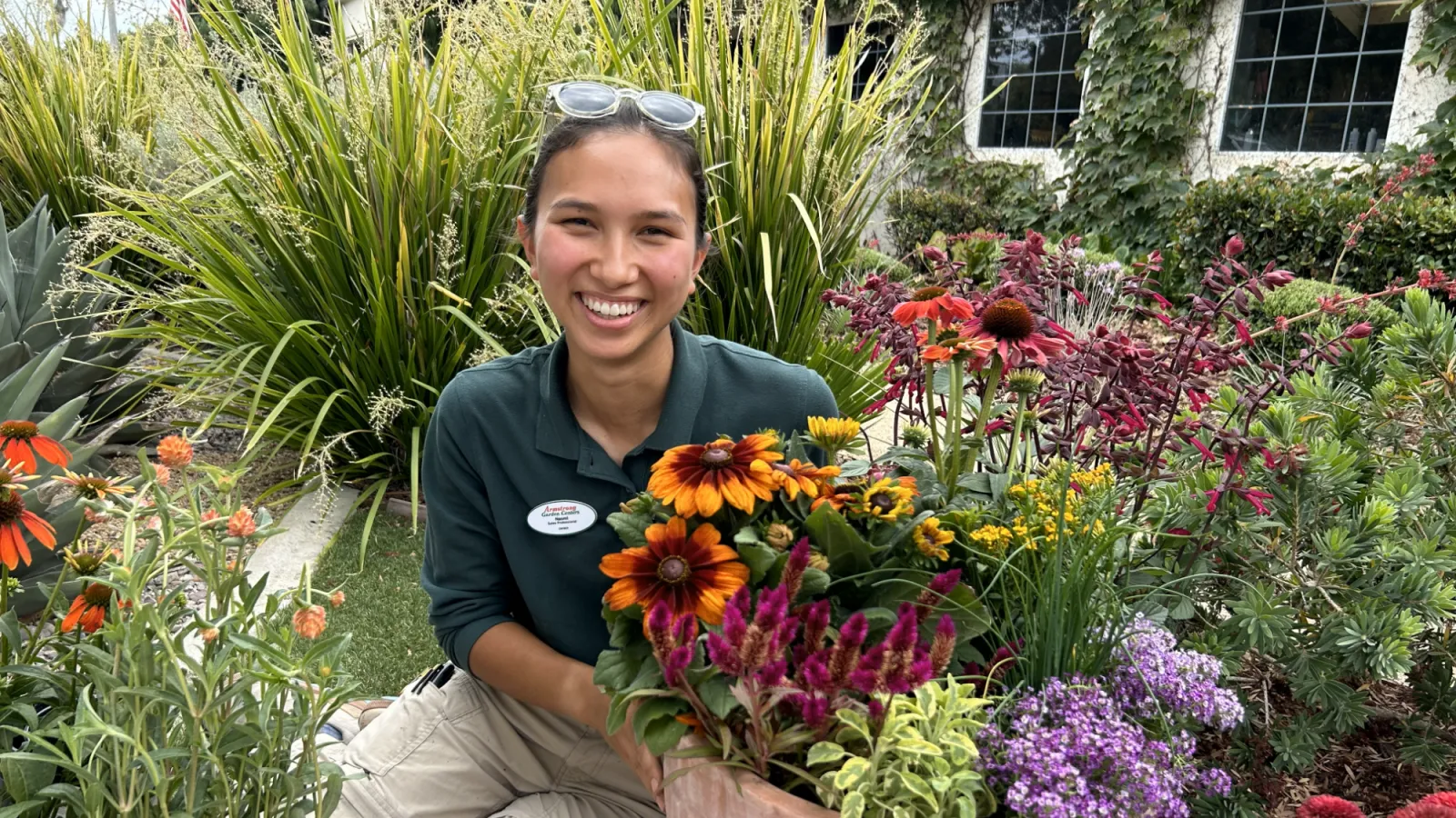 Smiling gardener with sunglasses on head holding a planter of colorful flowers in a vibrant garden setting.